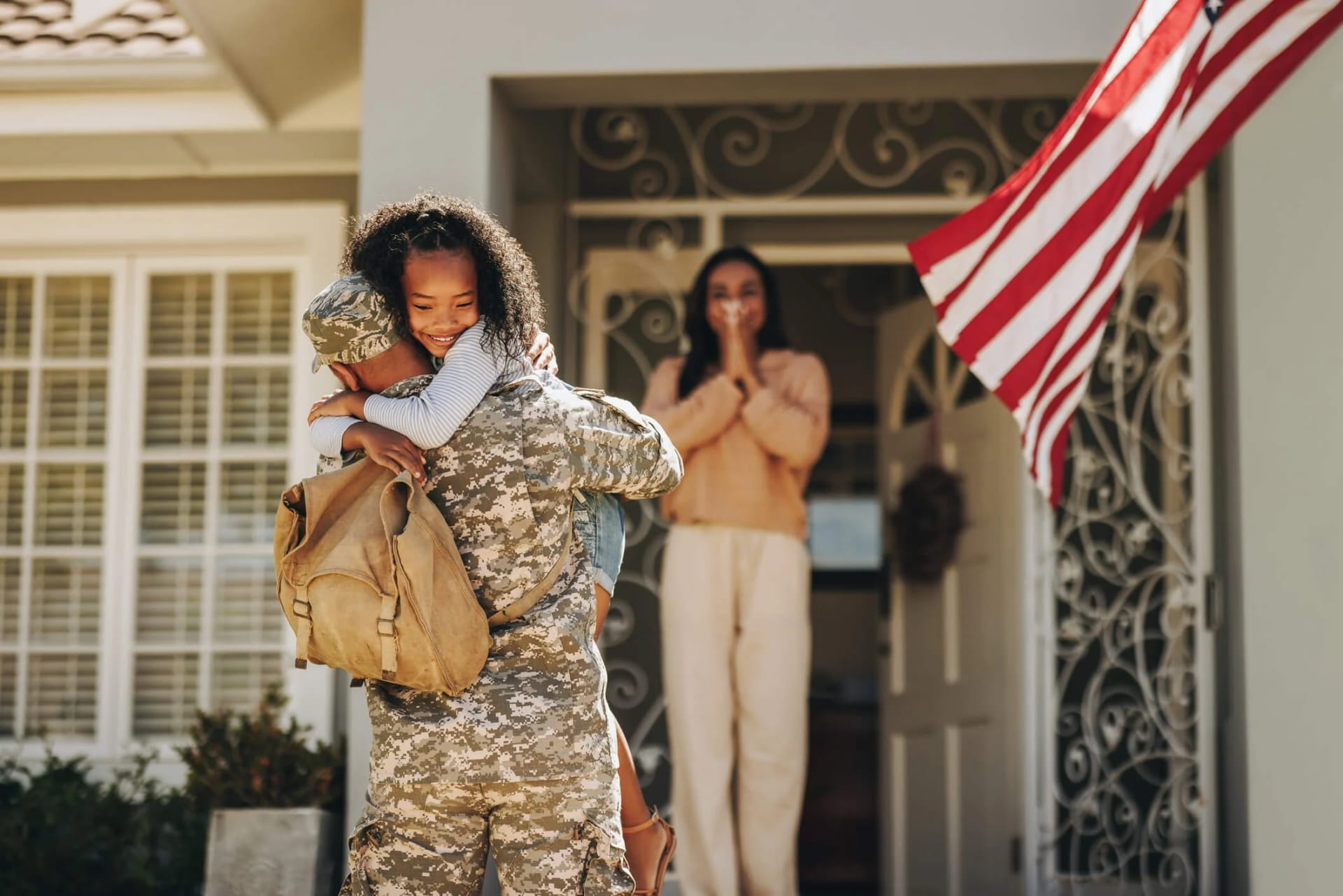 A service member in uniform hugs a child outside a family home as a woman watches from the doorway